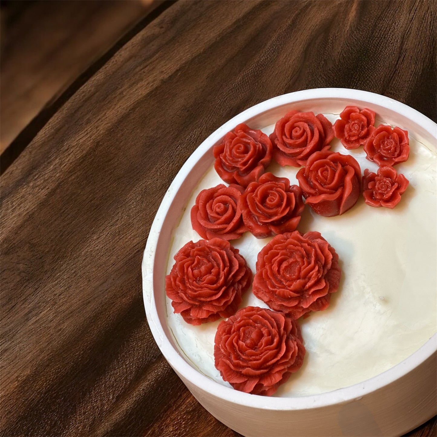Close-up, high-angle view of a round white ceramic dish candle containing deep red (burgundy) rose-shaped wax embeds, set against a dark, rustic wood background.