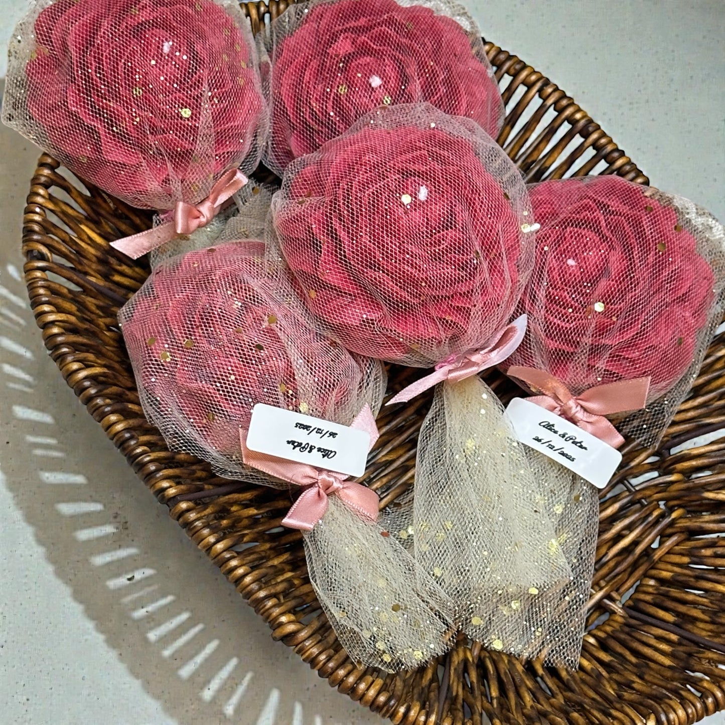 A rustic woven wicker basket filled with five deep pink, rose-shaped scented candles. Each candle is individually wrapped in sheer, light pink mesh fabric with gold glitter accents, tied with a delicate satin ribbon bow. Two favors feature small personalized tags with handwritten details, perfectly presented as elegant wedding or party return gifts.