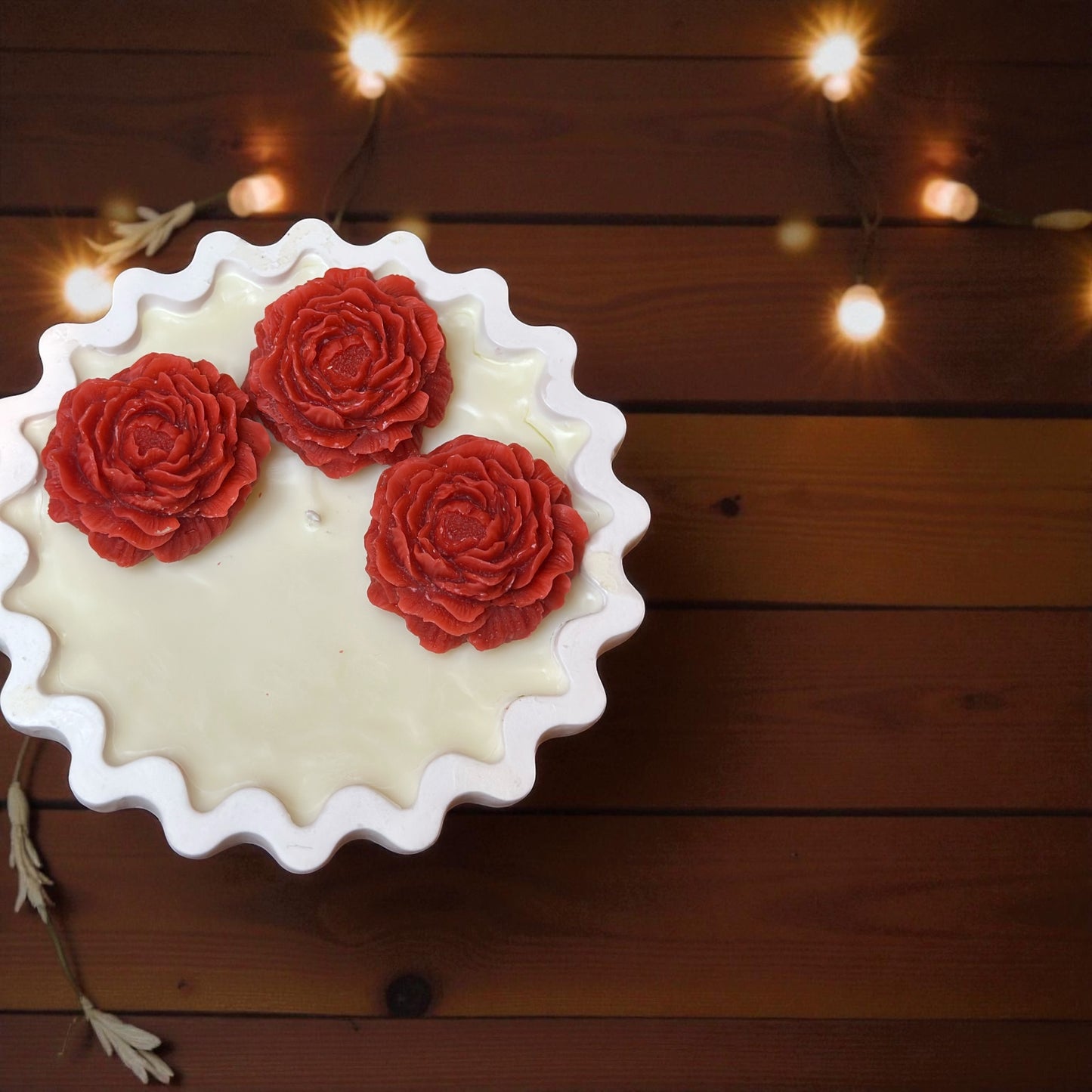 Unlit Scalloped Edge decorative candle with three large coral red wax roses. Hand-poured soy wax centerpiece on a dark wood grain background.