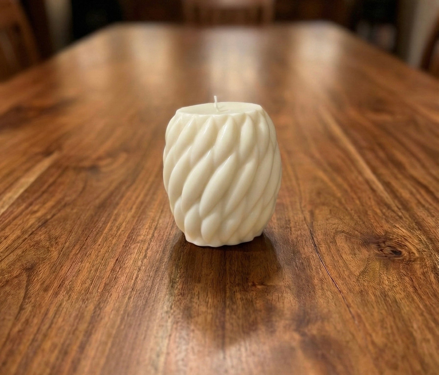 A cream-colored, twisted spiral pillar candle sitting on a wooden dining table. The candle features a ribbed, sculptural texture and is unlit, with a blurred warm home background