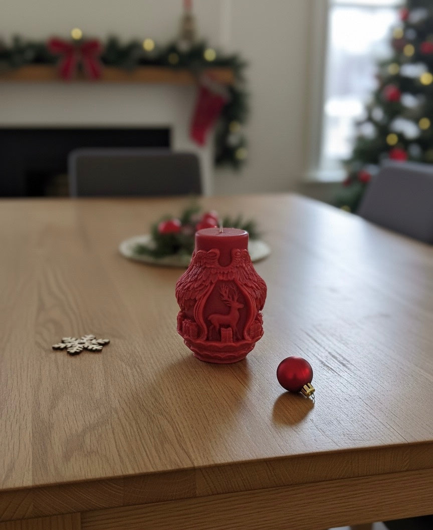 A detailed deep red sculptural candle shaped like angel wings wrapping around a standing reindeer. The candle sits on a wooden dining table with a blurred Christmas tree and fireplace garland in the background. A small wooden snowflake ornament lies next to it.