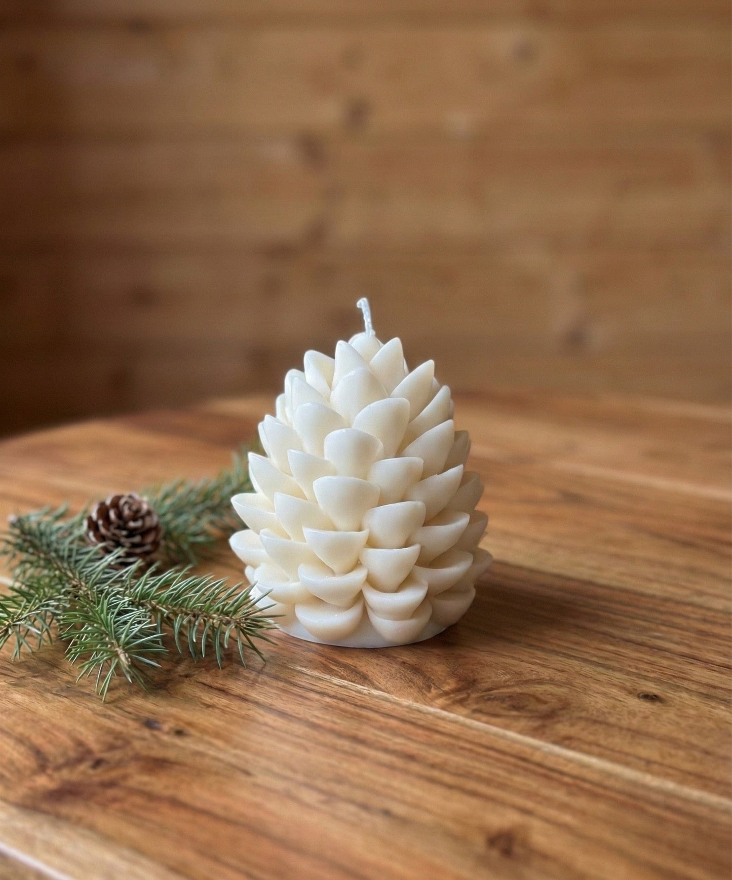 A single, cream-white pinecone shaped candle sitting on a wooden table. The candle is styled next to fresh green pine sprigs and a small real pinecone, creating a rustic Christmas look.