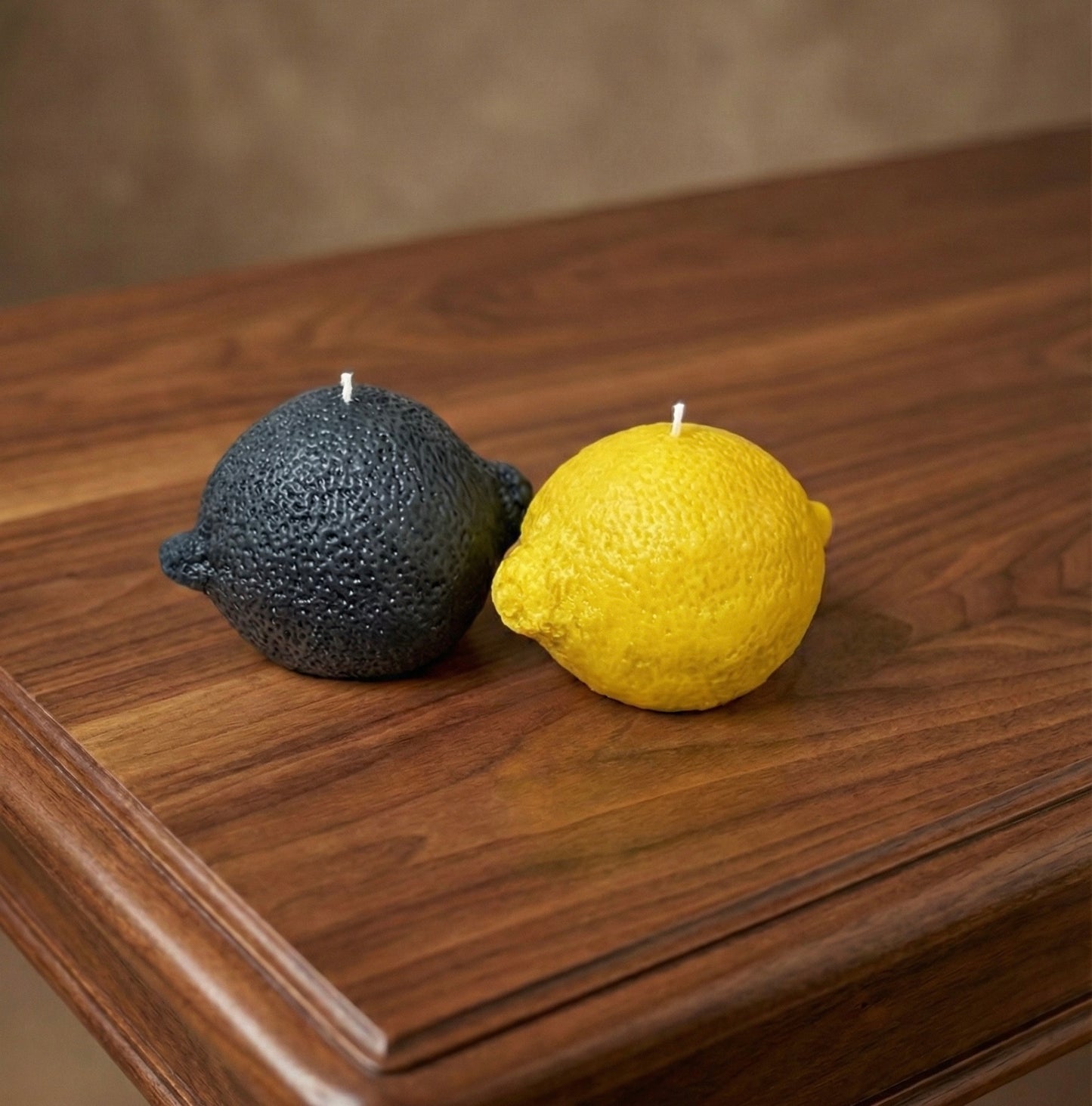 Two realistic lemon-shaped candles sitting on a polished dark wood table. On the left is a matte black lemon candle, and on the right is a vibrant yellow lemon candle. Both feature a textured, zest-like surface and white wicks.