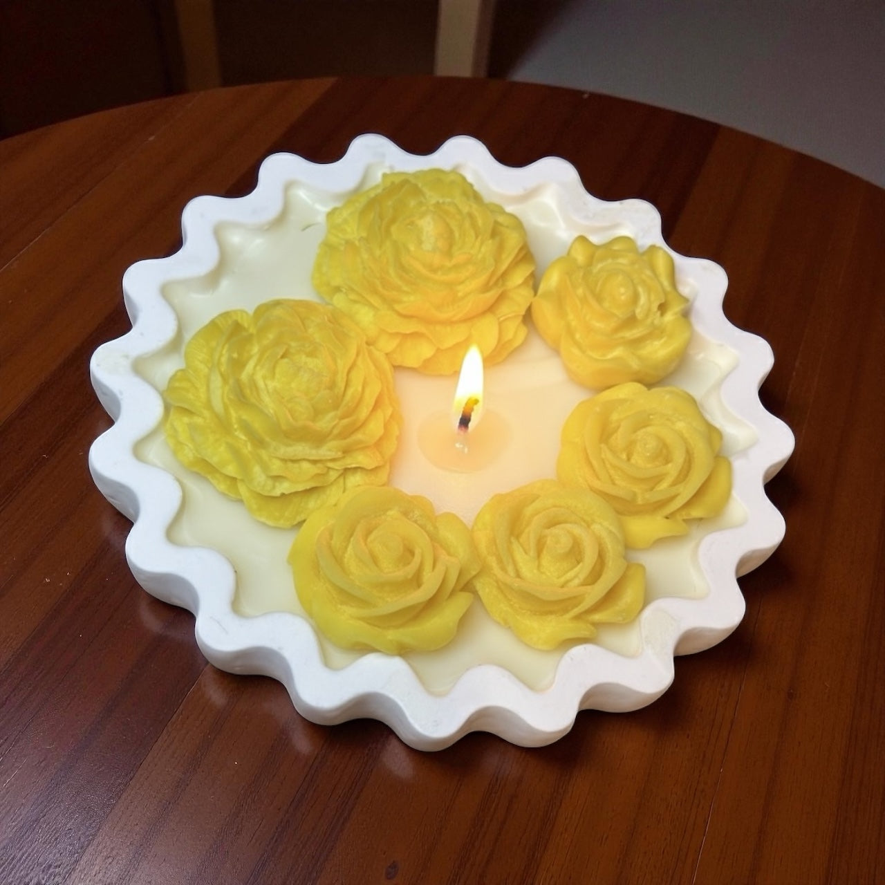 Lit Scalloped Edge decorative candle with an asymmetrical grouping of bright yellow wax roses and dahlias. Floral soy wax centerpiece on a wooden windowsill."