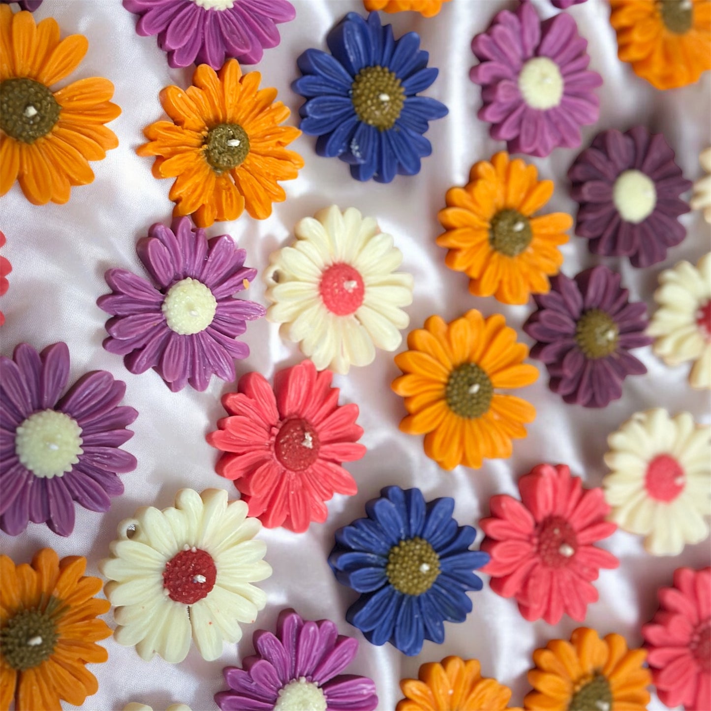 Overhead view of various colorful gerbera sunflower wax melts in shades of orange, blue, pink, and cream, scattered across a luxurious white silk fabric.