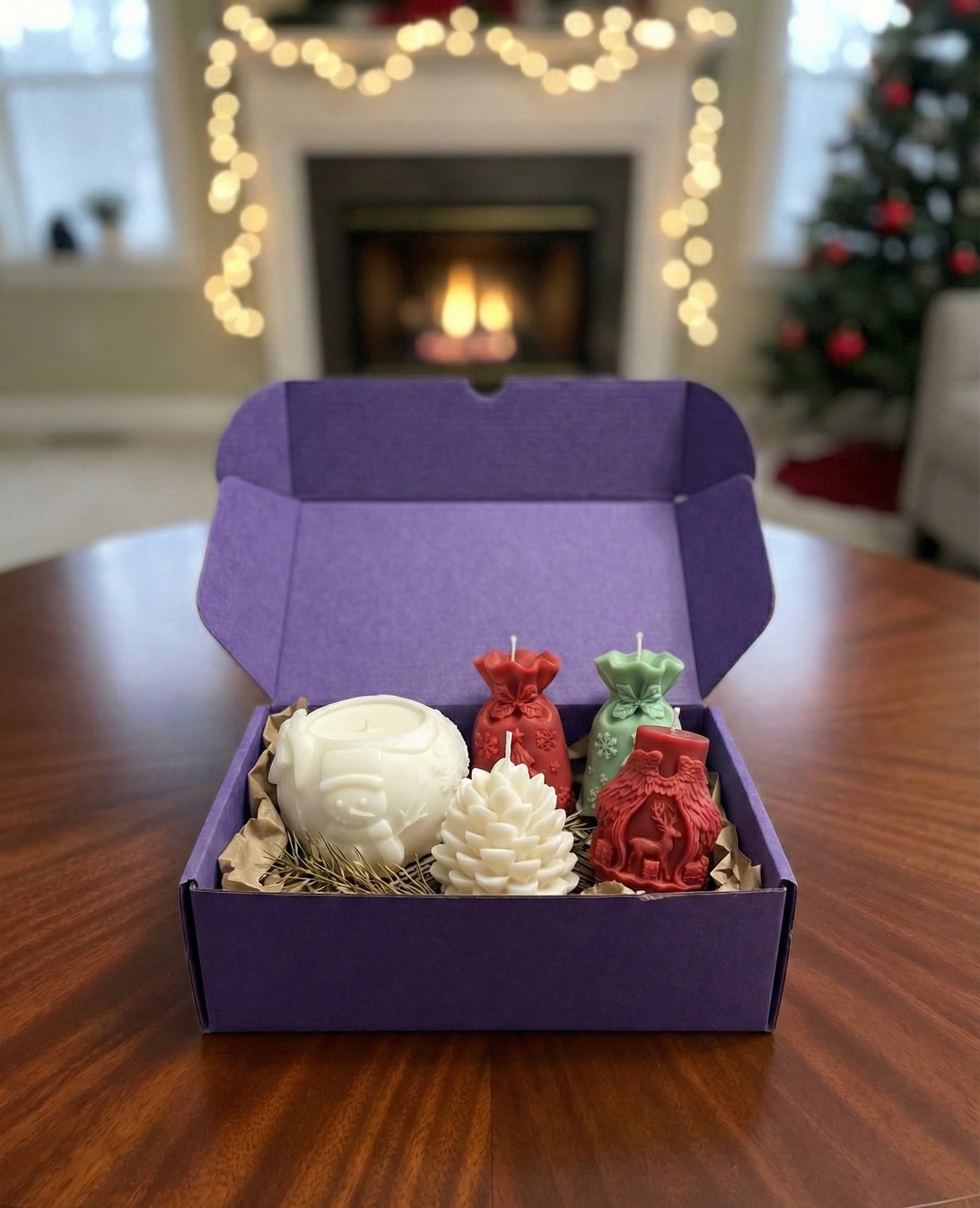 An open purple gift box displayed on a table with a cozy, blurred fireplace and Christmas tree in the background. The box contains five festive candles: a white snowman bowl, a white pinecone, red and green gift sack candles, and a red ornate pillar candle with a reindeer design, nestled in brown crinkle paper.