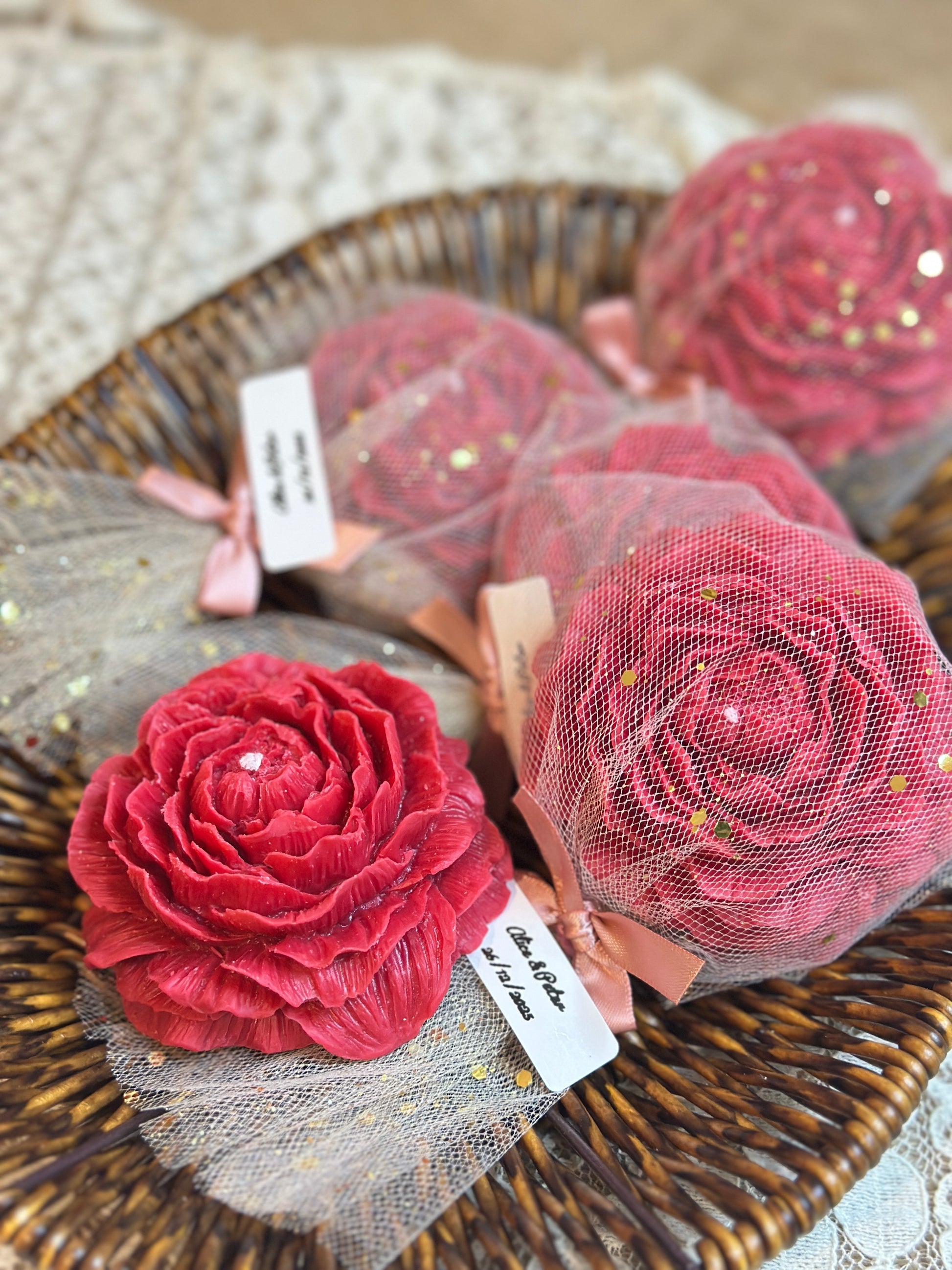 A close-up view of a deep pink, realistic rose bloom scented candle resting on gold-flecked sheer mesh fabric, next to a wicker basket. The basket holds four more matching rose candles, wrapped and tied with pink ribbon. The texture of the candle's petals and the glitter in the packaging are highlighted, emphasizing the product's quality as a luxurious personalized party favor.