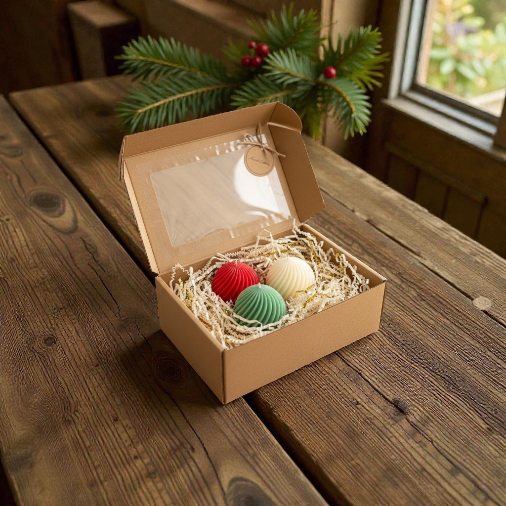 Christmas gift set of three festive swirl ball candles (red, green, white) packaged in an open brown cardboard box with a clear window, on a rustic wooden table with pine branches and red berries in the background.