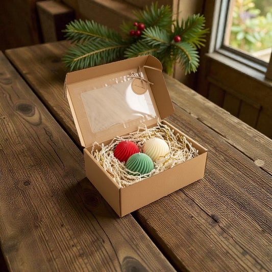 Christmas gift set of three festive swirl ball candles (red, green, white) packaged in an open brown cardboard box with a clear window, on a rustic wooden table with pine branches and red berries in the background.