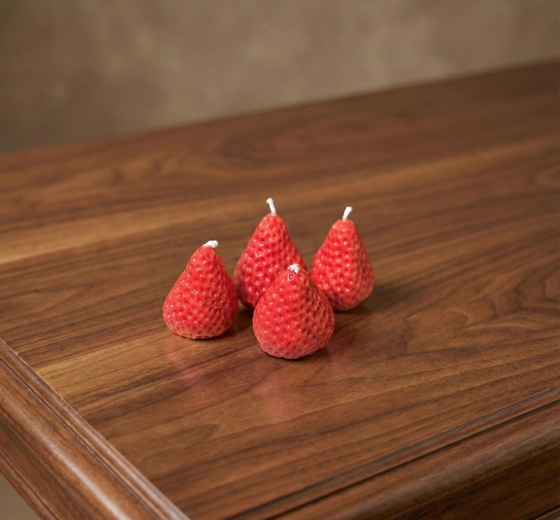 Four realistic, red strawberry-shaped candles with textured seeds and white wicks arranged on a polished dark wood table.