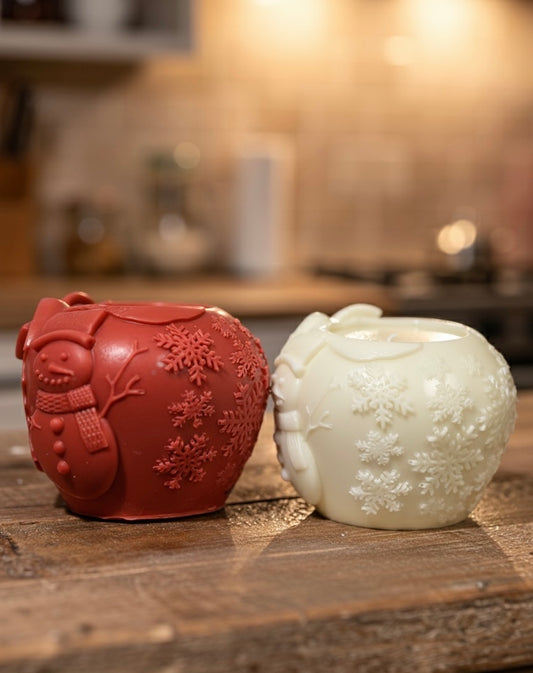 Comparison view of a red silicone mold next to a finished white wax sphere candle featuring an embossed snowman and snowflake design on a wooden surface