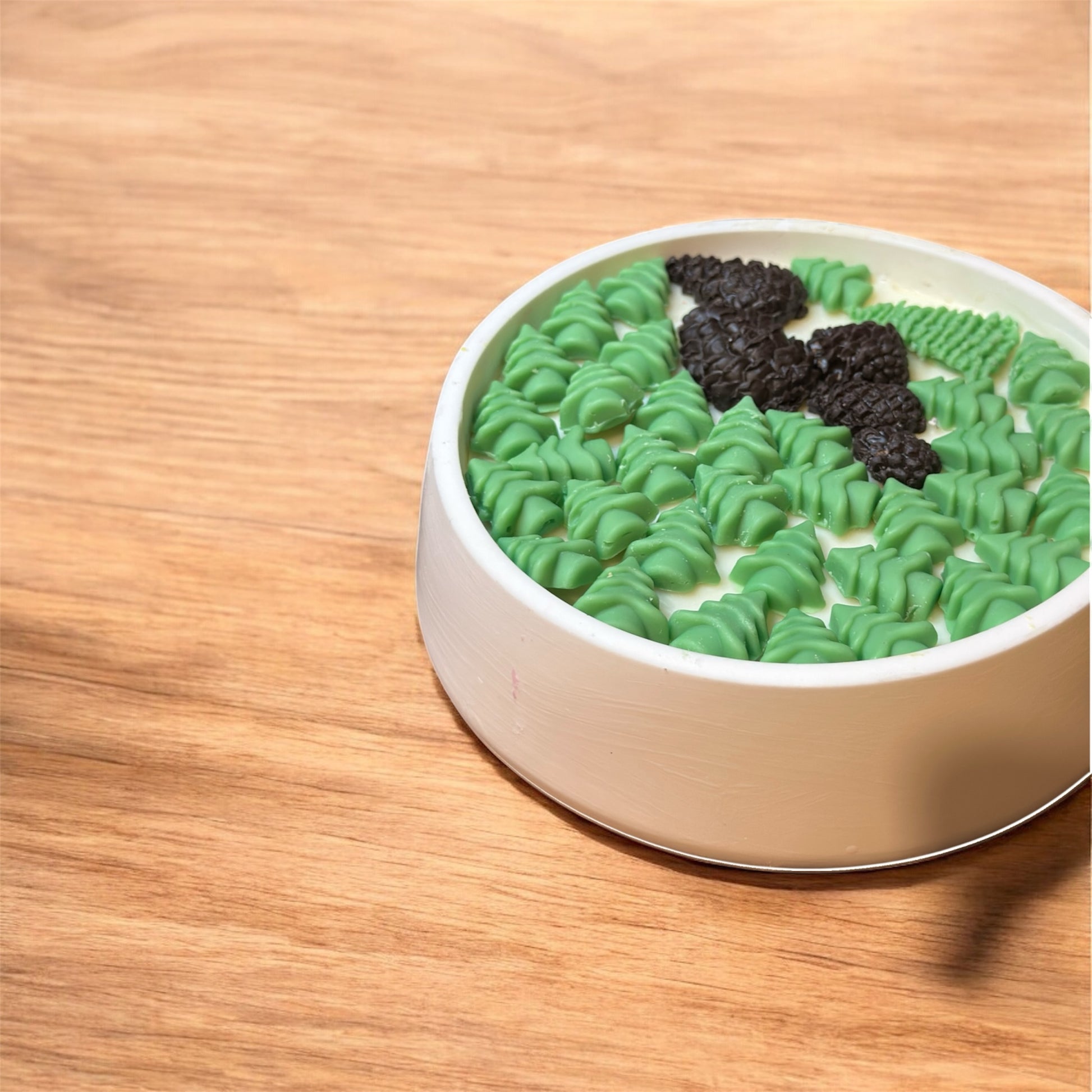 Top-down view of a round white candle with green tree and dark brown pinecone-shaped wax melts floating on top, against a light wooden table.