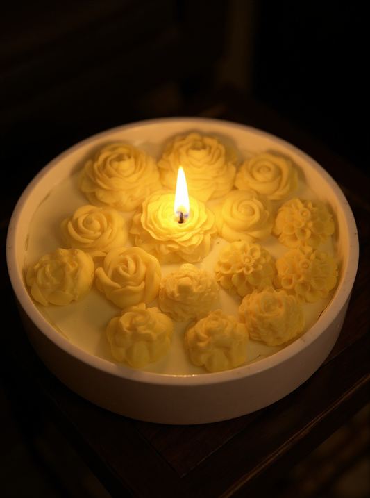 A wide, round white ceramic bowl candle filled with bright yellow rose and dahlia wax embeds, lit with two wicks, sitting on a wooden surface with soft sunlight.