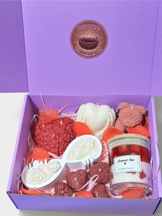 A purple gift box filled with various handcrafted Valentine's Day candles including a red floral heart, a white infinity symbol candle with small roses, a rose-textured teddy bear, and a glass jar candle labeled "Forever You." The box is decorated with pink paper shredding and red rose petals.