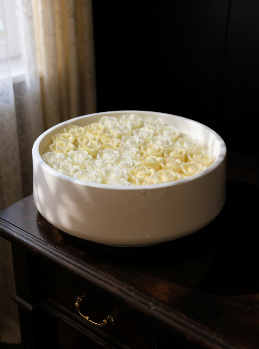 Close-up of a large round ceramic candle featuring a pattern of small white and pale yellow wax roses, set next to a steaming glass of tea on dark wooden furniture in natural daylight