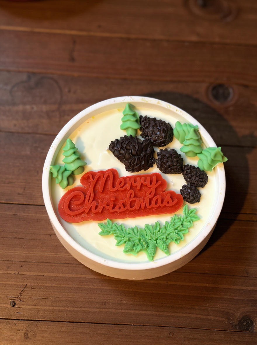 Overhead shot of a decorative round white Christmas candle, with a vibrant red 'Merry Christmas' message, assorted green wax trees and branches, and dark brown wax pinecones arranged artistically on top of the creamy candle wax