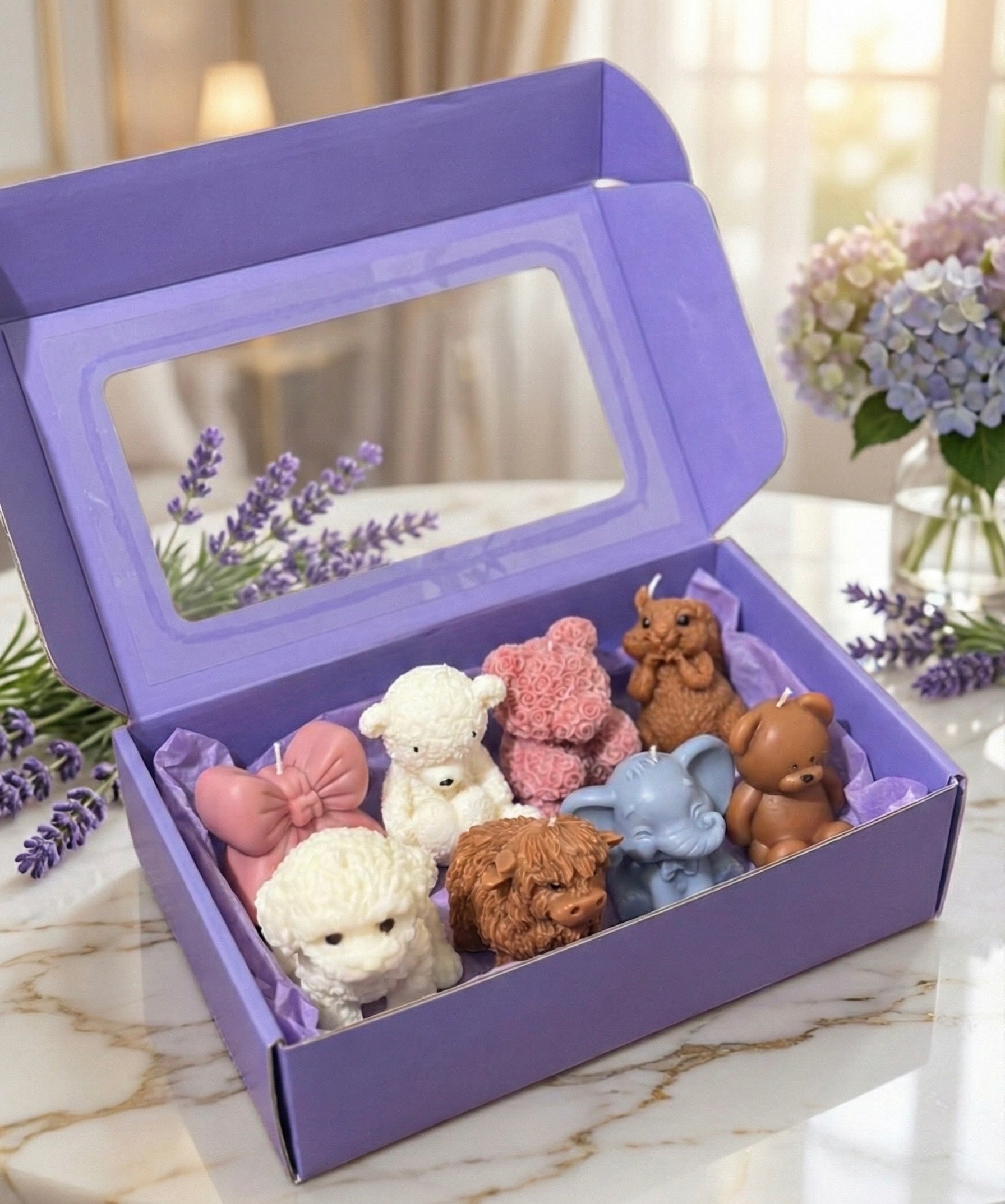 Large, open purple gift box with a clear window on the lid. The box contains eight assorted shaped candles including a highland cow, white and brown teddy bears, a pink rose bear, an elephant, and a pink bow. Lavender flowers are in the background.