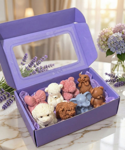Large, open purple gift box with a clear window on the lid. The box contains eight assorted shaped candles including a highland cow, white and brown teddy bears, a pink rose bear, an elephant, and a pink bow. Lavender flowers are in the background.