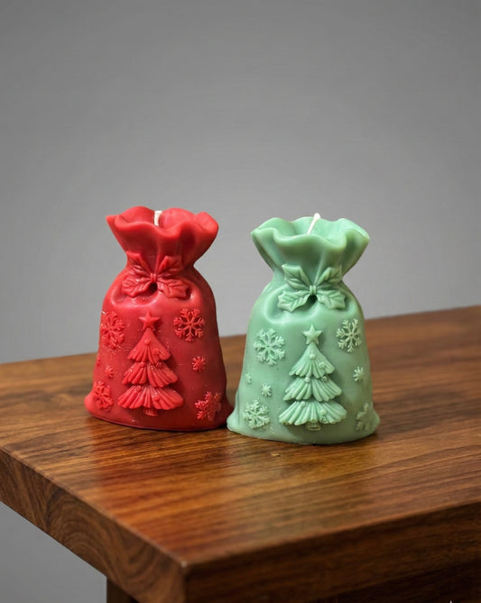 Two sculptural Christmas candles shaped like Santa's gift sacks sitting on a wooden table. One is deep red and the other is soft green. Both feature embossed designs of a Christmas tree with a star topper and scattered snowflak