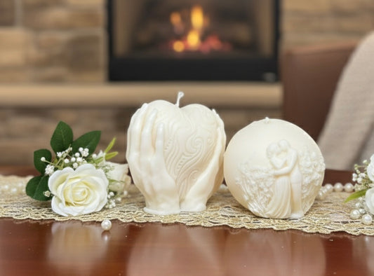Two white sculptural candles on a gold lace runner: one shaped like hands holding a textured heart and another spherical candle featuring a carved embracing couple.
