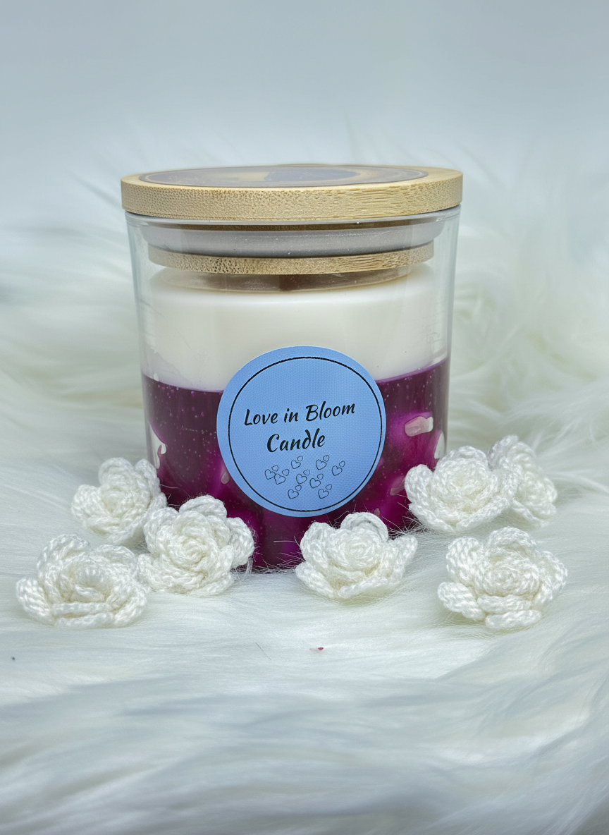 A two-layered candle in a glass jar with a bamboo lid, featuring a white top layer and a purple bottom layer. The label reads "Love in Bloom Candle." The jar is surrounded by small white crocheted roses on a white faux fur surface.