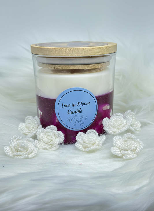 A two-layered candle in a glass jar with a bamboo lid, featuring a white top layer and a purple bottom layer. The label reads "Love in Bloom Candle." The jar is surrounded by small white crocheted roses on a white faux fur surface.