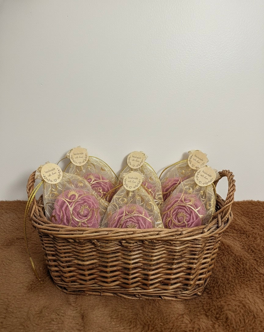 A wicker basket filled with multiple pink rose party favors, each individually wrapped in a gold and white organza drawstring pouch. The basket of bulk return gifts is set against a white wall on a brown fur surface, showcasing the quantity and presentation.