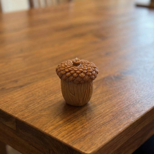 Close-up of a brown, acorn-shaped decorative candle sitting on a wooden table. The candle features a highly textured, bumpy cap and a base with vertical wood-grain carving details, perfect for rustic autumn decor.