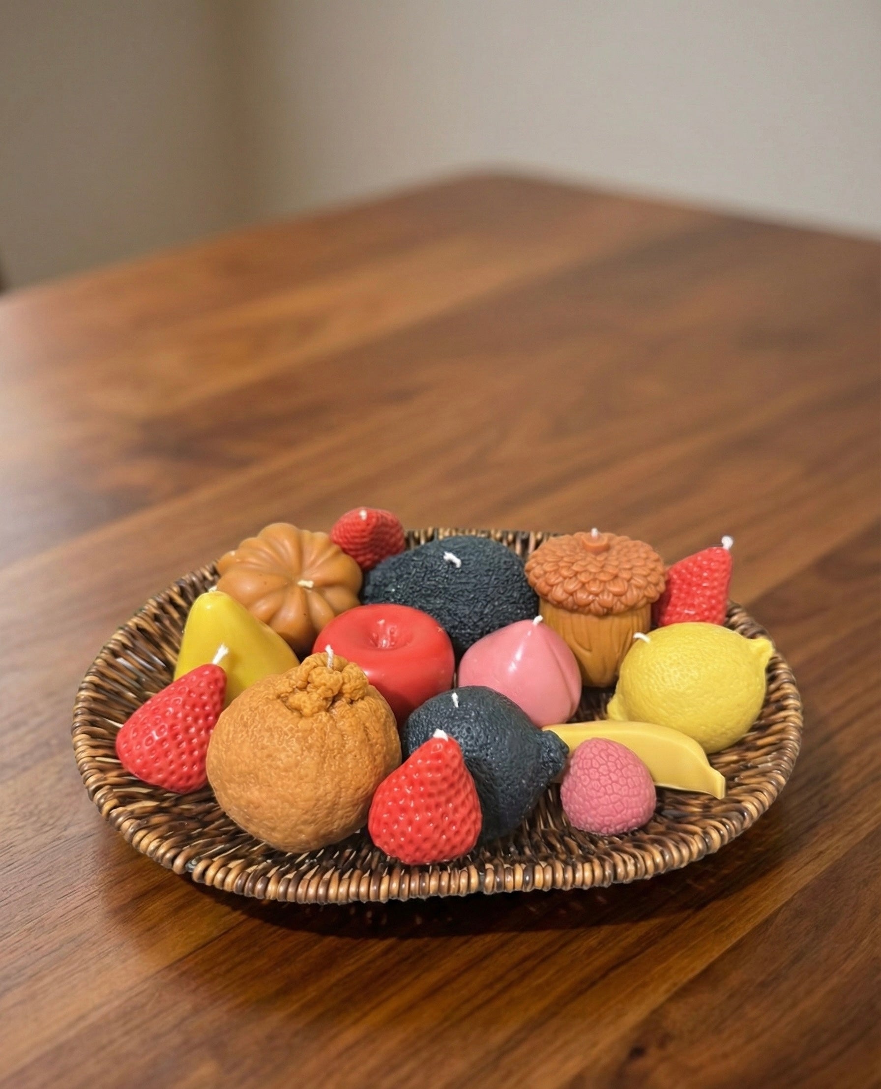 A close-up side view of a woven wicker plate resting on a wooden table, filled with an assortment of realistic fruit-shaped candles, including strawberries, an avocado, a lemon, and an apple.