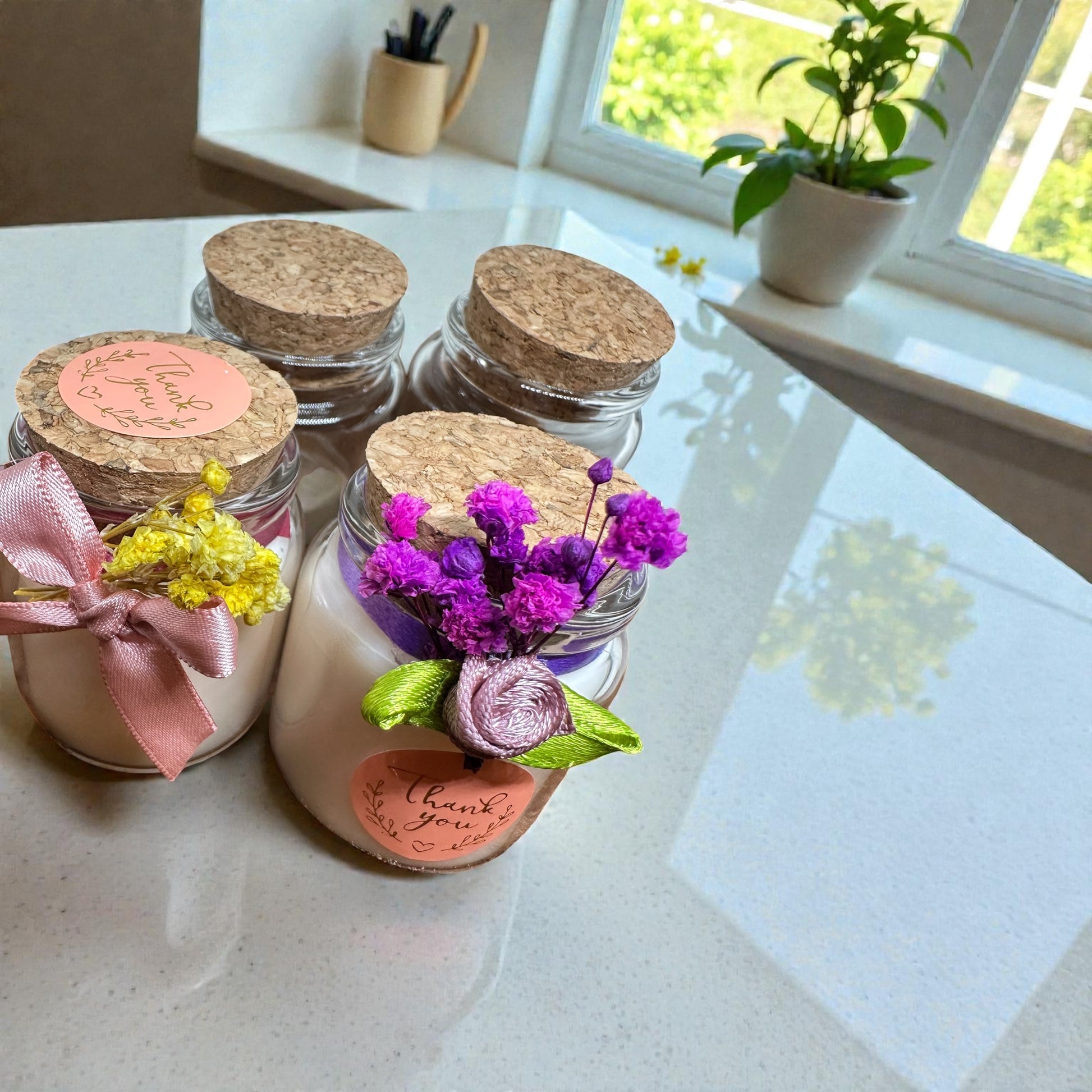 Close-up of four mini glass jar candles, showing two different styles: one with a pink ribbon and yellow flowers, and one with a purple ribbon and purple flowers. They have cork lids and "Thank You" stickers, set on a bright kitchen counter. 50ml personalized favor candles.