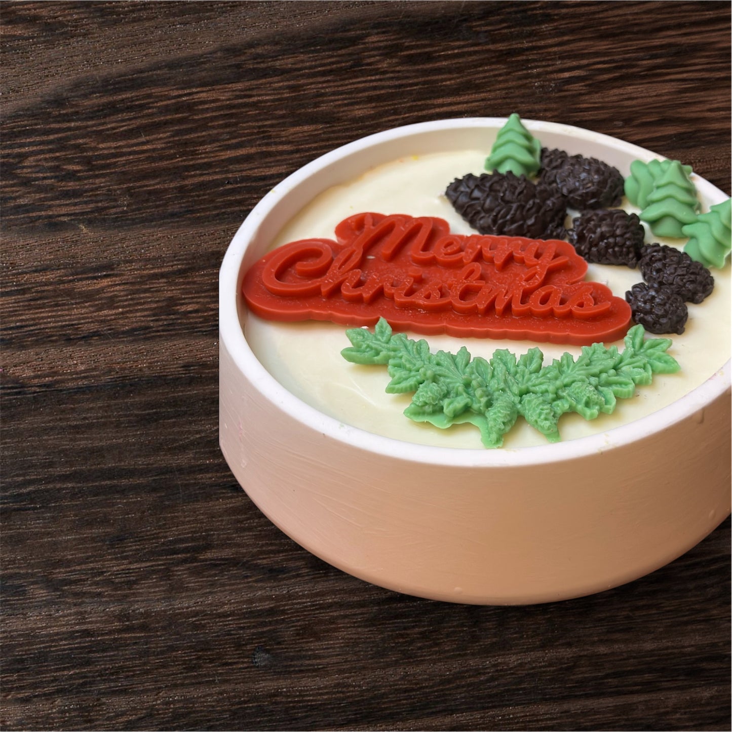 Angled view of a festive round white candle, showcasing a prominent red 'Merry Christmas' wax centerpiece surrounded by small green wax trees, pine branches, and dark brown wax pinecones, set on a warm wooden surface