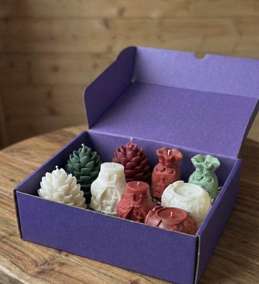 An open, deep purple gift box sitting on a wooden table, filled with a large assortment of Christmas-themed shaped candles. The set includes white, sage green, and berry red candles shaped like pinecones, gift sacks with bows, and ornate holiday jars featuring reindeer and angel designs.