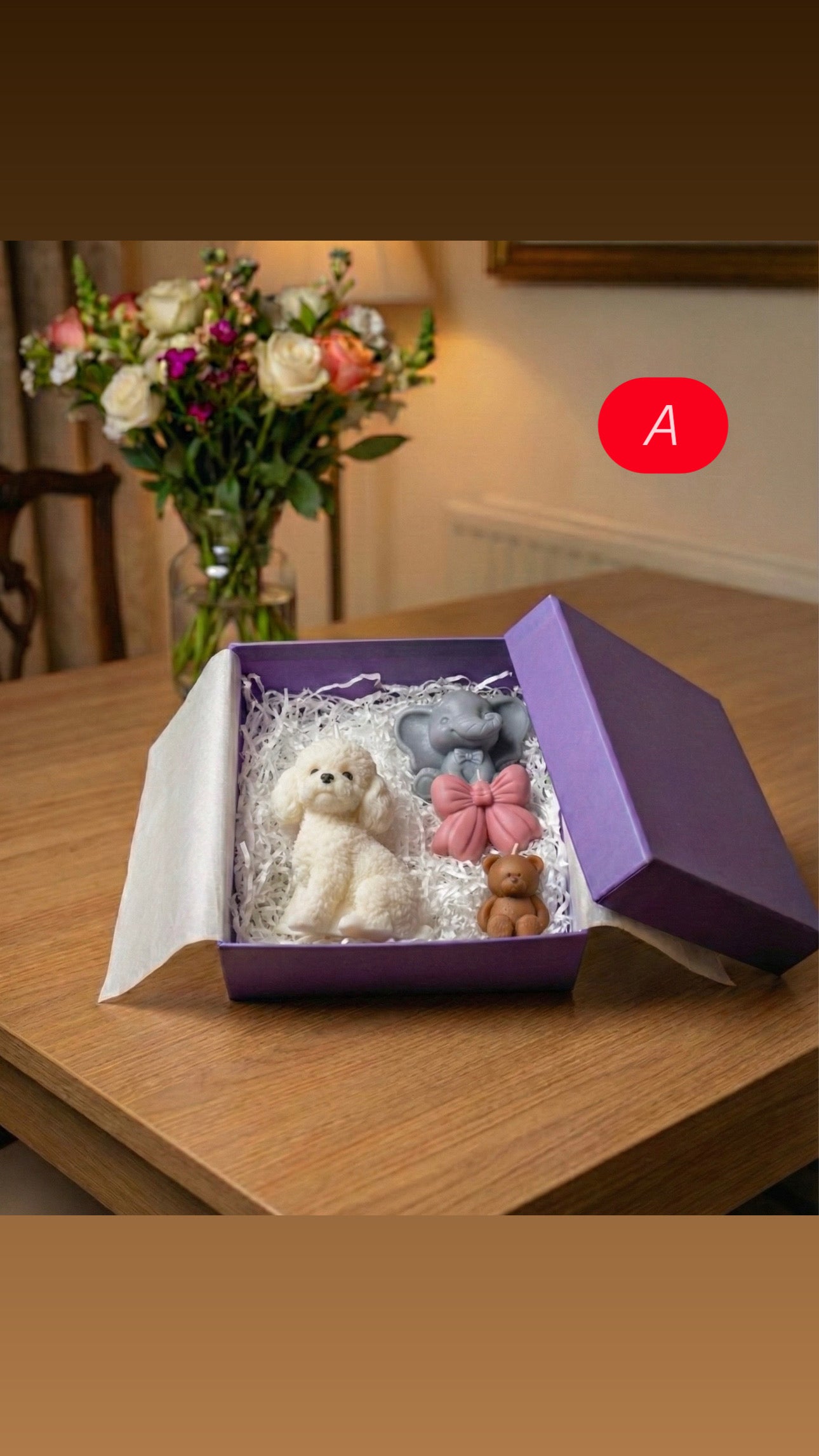 Open purple gift box labeled A sitting on a wooden table. Inside are four shaped candles: a white poodle dog, a grey elephant, a decorative pink bow, and a small brown teddy bear, nestled in white shredded paper.