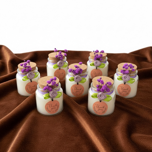 Seven miniature scented candles in clear glass jars with cork lids, arranged on a brown velvet cloth. Each 50ml party favor candle is decorated with a purple ribbon, a small orange-pink "Thank you" sticker, and a bunch of lilac dried flowers. Excellent as baby shower or christening return gifts.