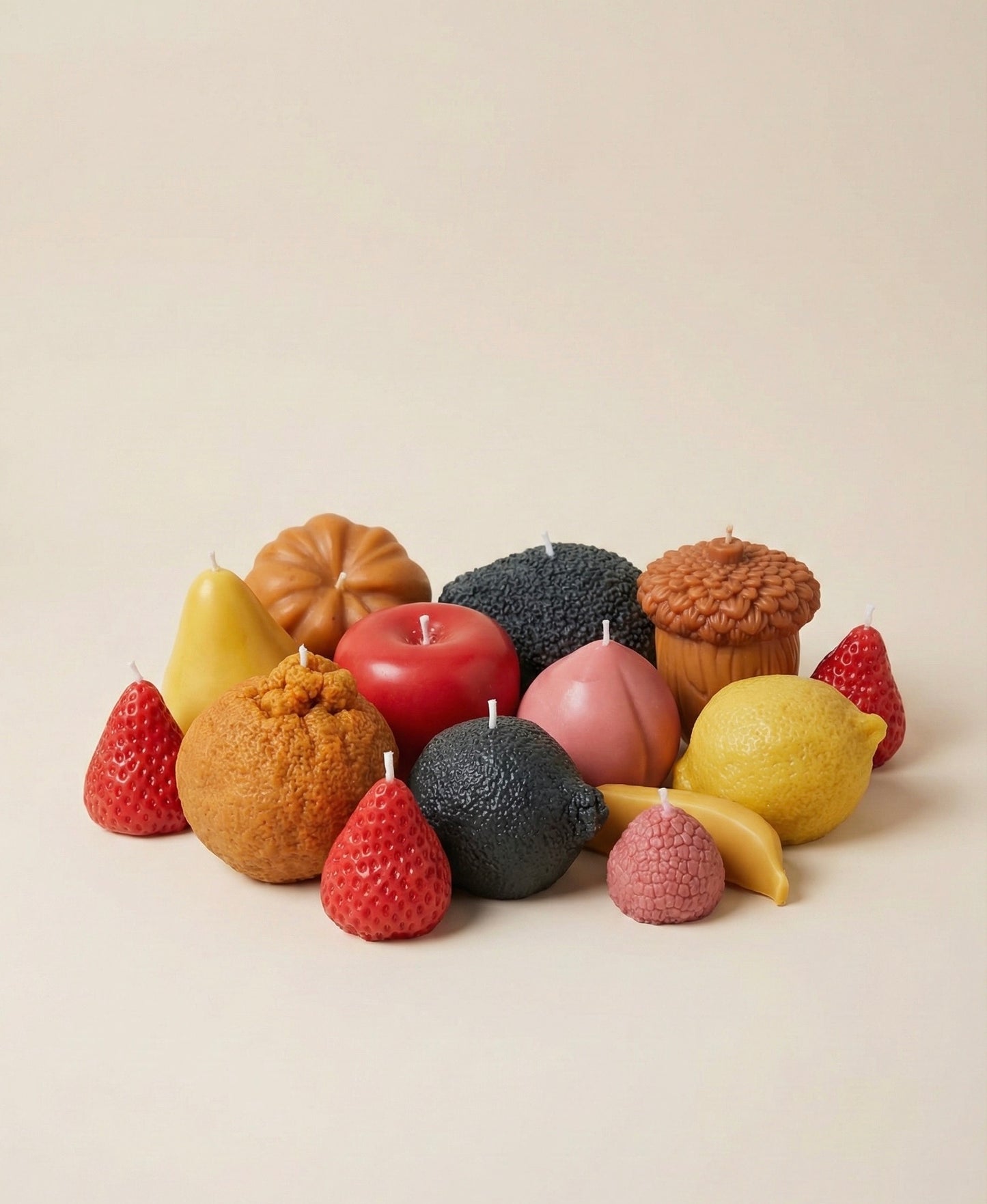 A studio shot of 13 colorful, novelty wax candles shaped like various fruits arranged on a plain beige background. The group includes a pear, peach, acorn, banana, and textured citrus fruits.