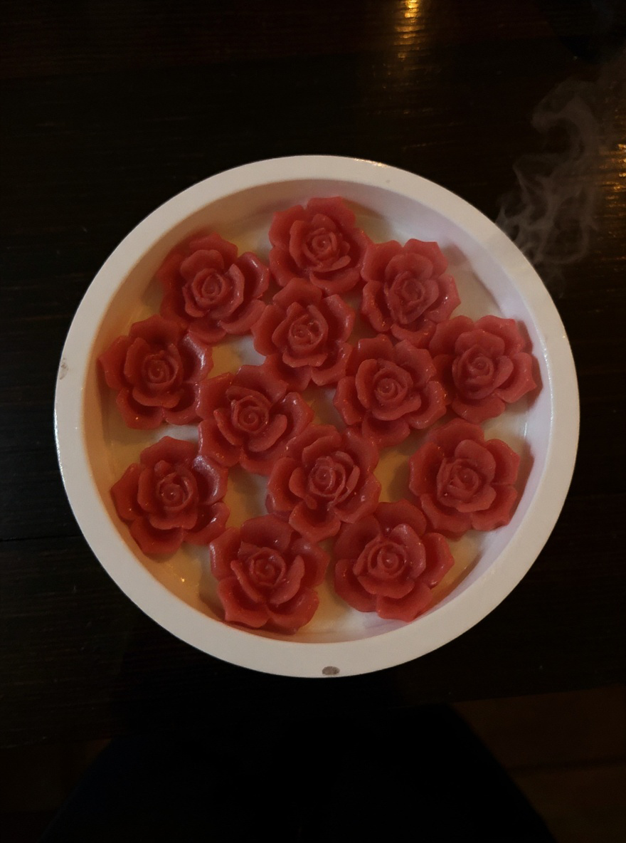 Vibrant Coral Red Rose decorative candle in a white round ceramic bowl. Hand-poured soy wax styled next to two small amber glass apothecary bottles."