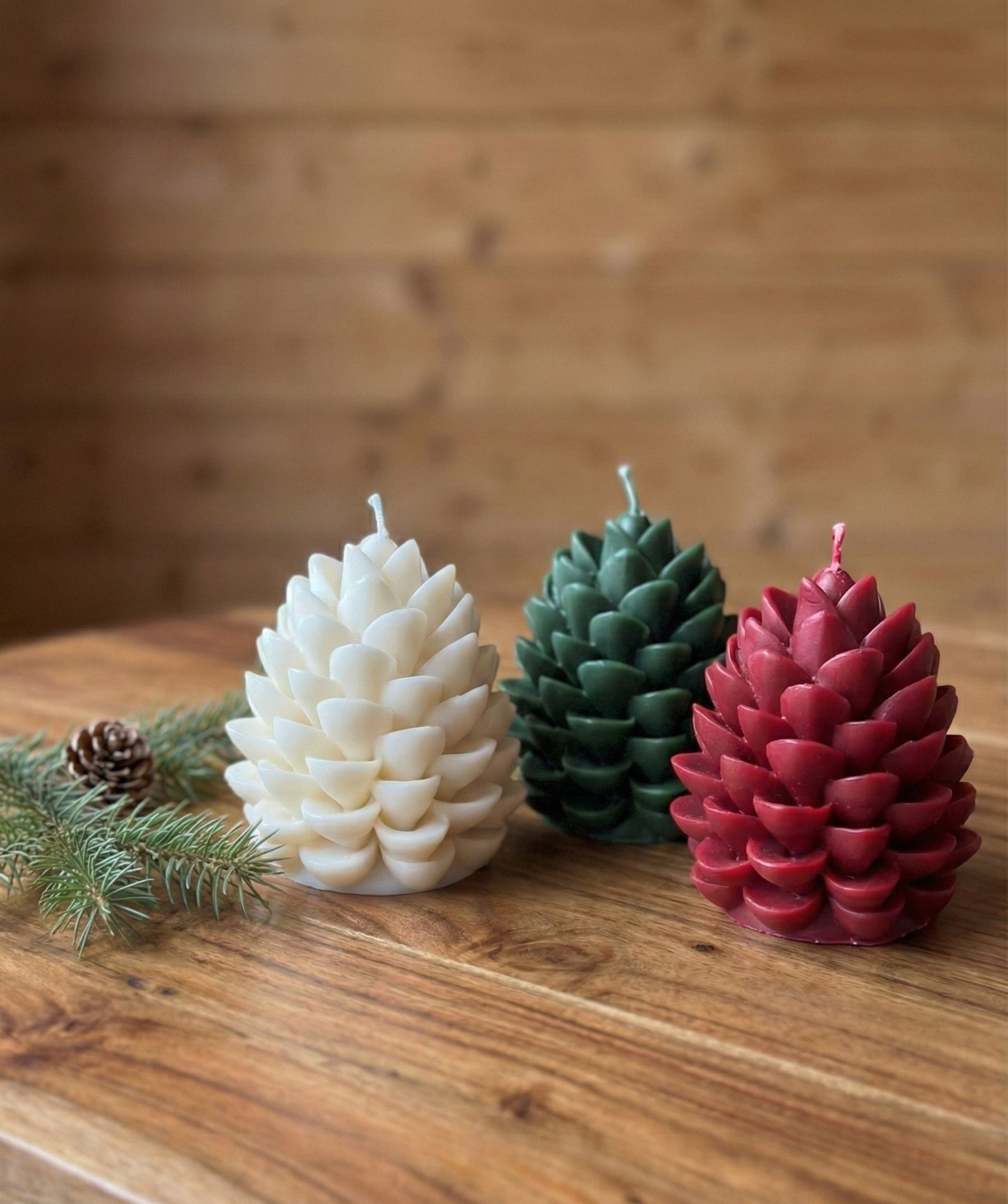 A trio of festive pinecone candles arranged on a wood surface. From left to right: a white candle, a forest green candle, and a deep red burgundy candle, styled with pine branches for holiday home decor.
