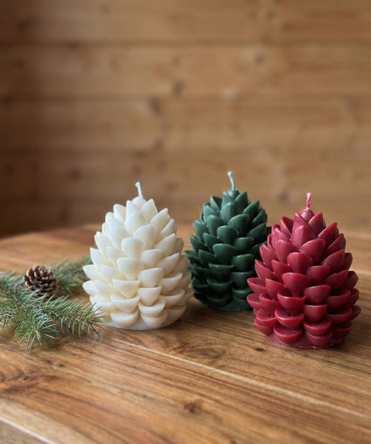 A trio of festive pinecone candles arranged on a wood surface. From left to right: a white candle, a forest green candle, and a deep red burgundy candle, styled with pine branches for holiday home decor.