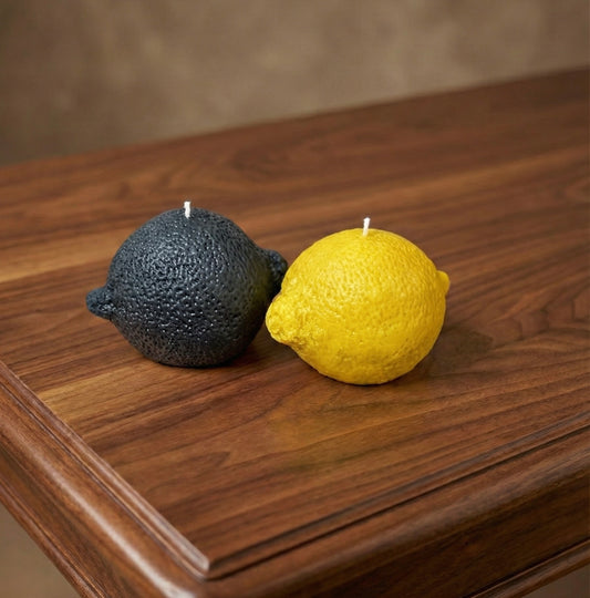 Two realistic lemon-shaped candles sitting on a polished dark wood table. On the left is a matte black lemon candle, and on the right is a vibrant yellow lemon candle. Both feature a textured, zest-like surface and white wicks.