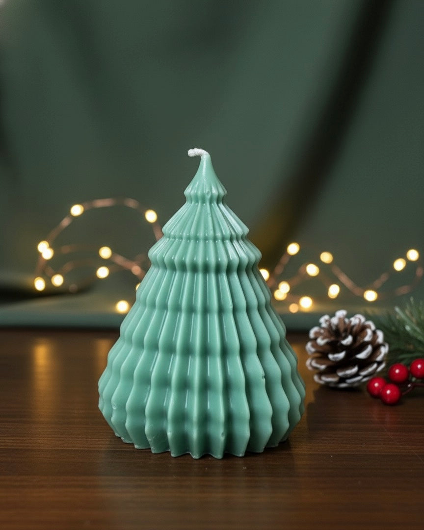 Large Christmas tree-shaped candle in a subtle sage green color, sitting on a wooden table with blurred fairy lights and a pinecone in the background