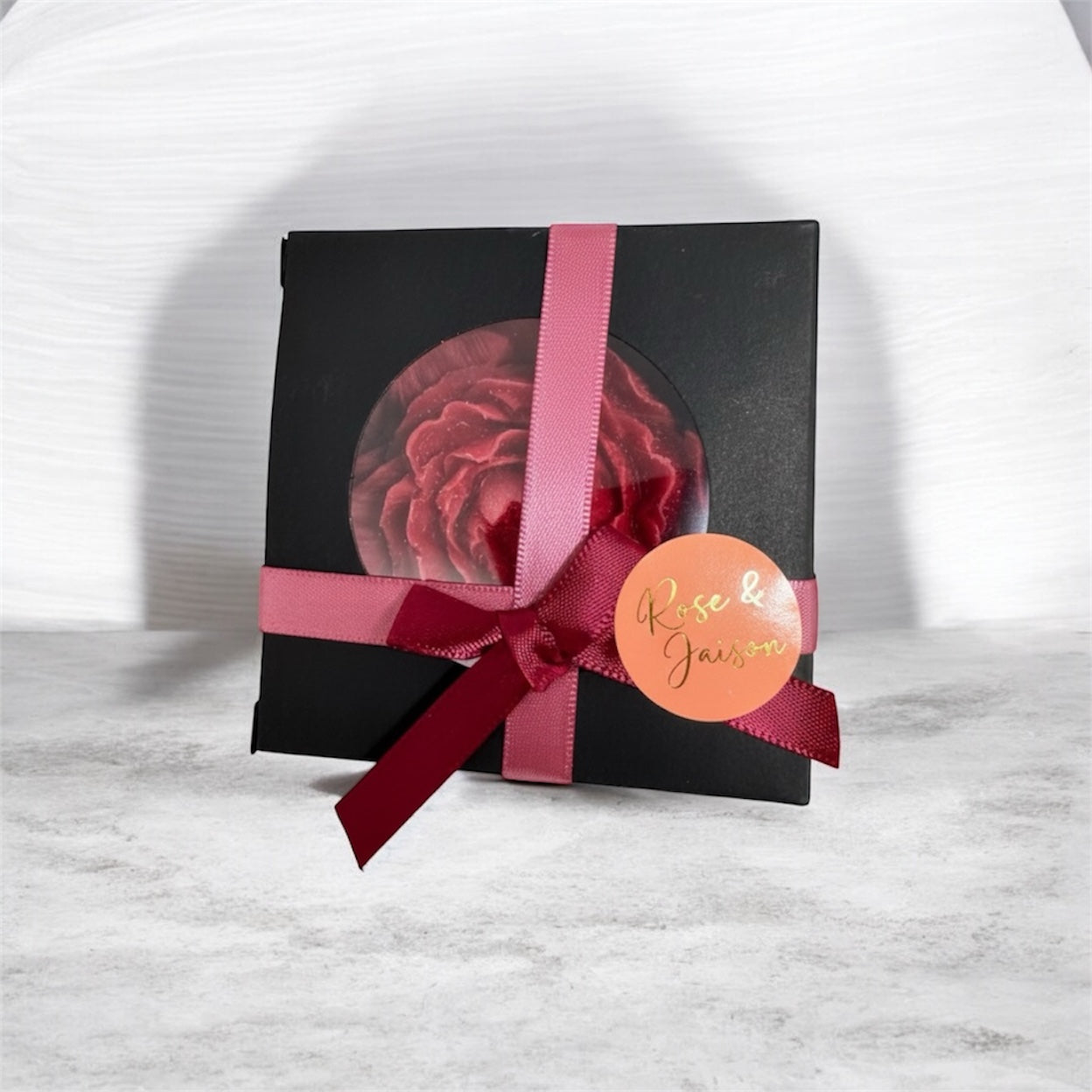Close-up of a single elegant red rose or peony bloom candle favor in a matte black box with a clear window. The box is cinched with a dusty rose and deep burgundy ribbon, featuring a prominent coral-colored circular tag with gold text that reads 'Rose & Jaison.' The favor is set against a white wood and gray marble-look background, showcasing the beautiful return gift packaging.