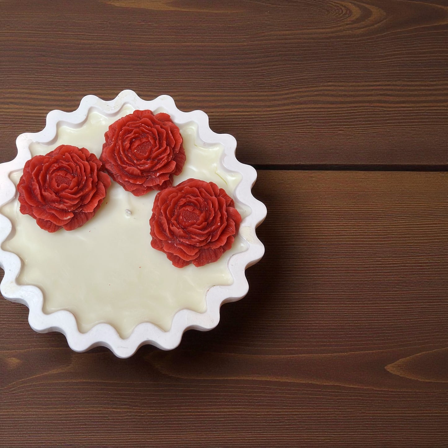 Unlit Scalloped Edge decorative candle with three large coral red wax roses. Hand-poured soy wax centerpiece on a dark wood grain background.