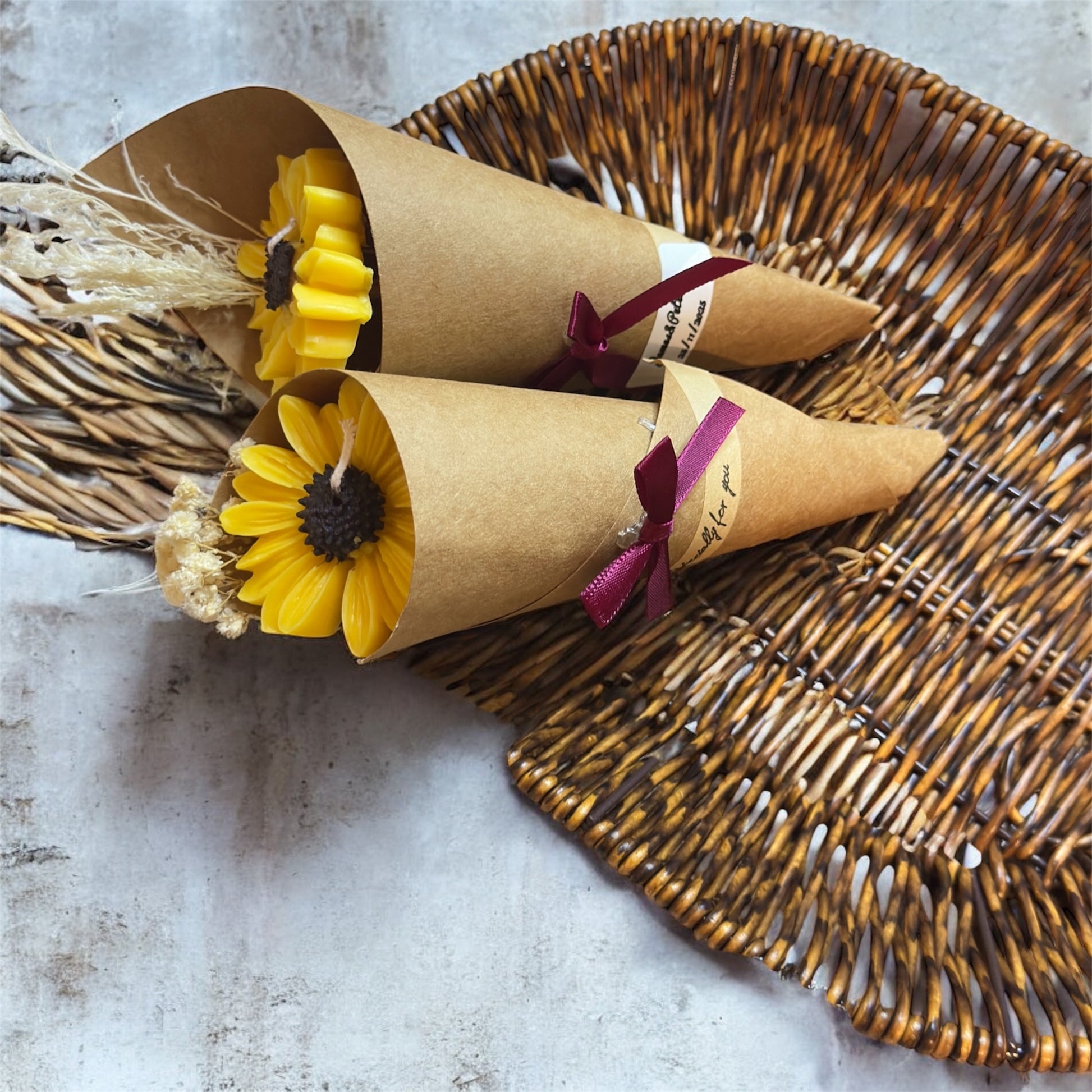 Two handmade yellow sunflower candles wrapped in brown kraft paper cones with burgundy ribbon bows, placed on a light brown woven wicker basket. They are perfect as rustic wedding favors