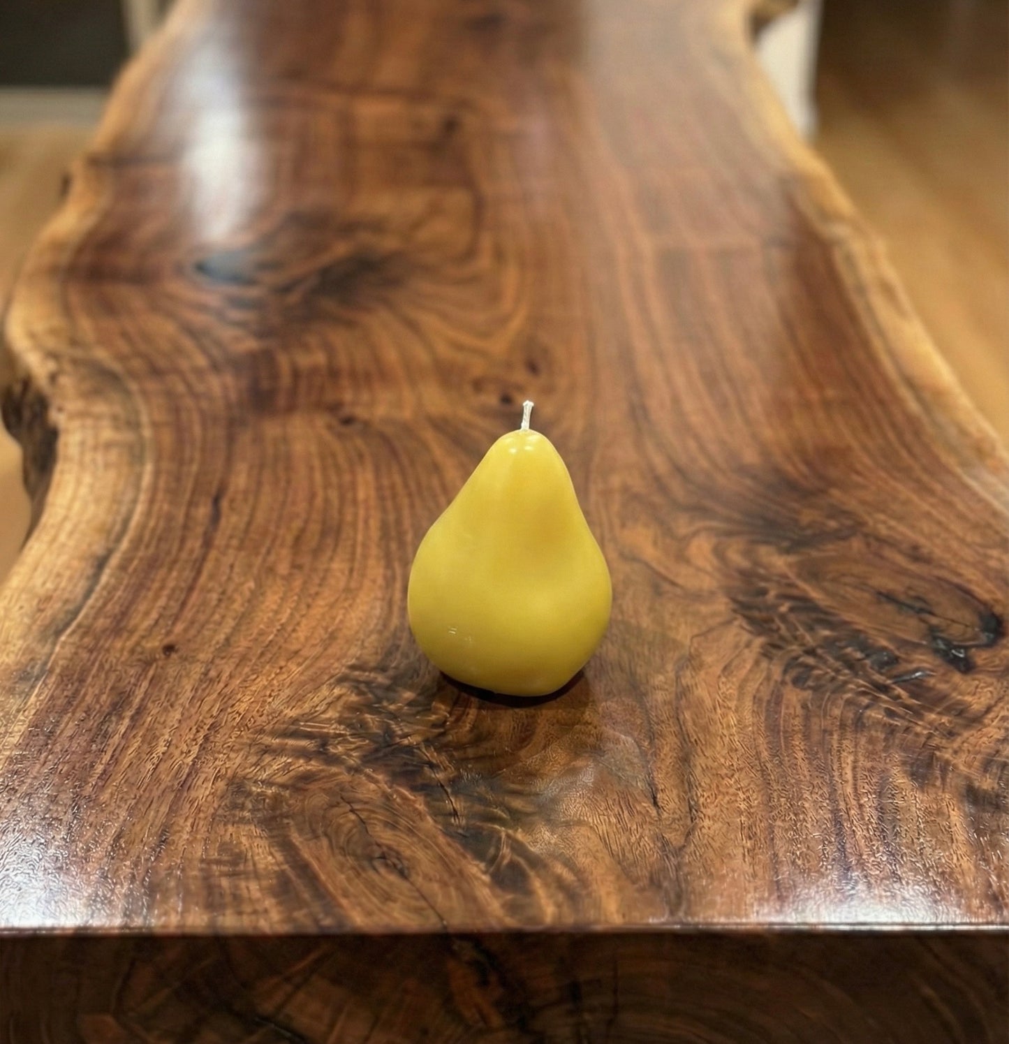 "A realistic, yellow pear-shaped beeswax candle with a white wick, sitting on a polished dark wood live-edge table with a blurred background."