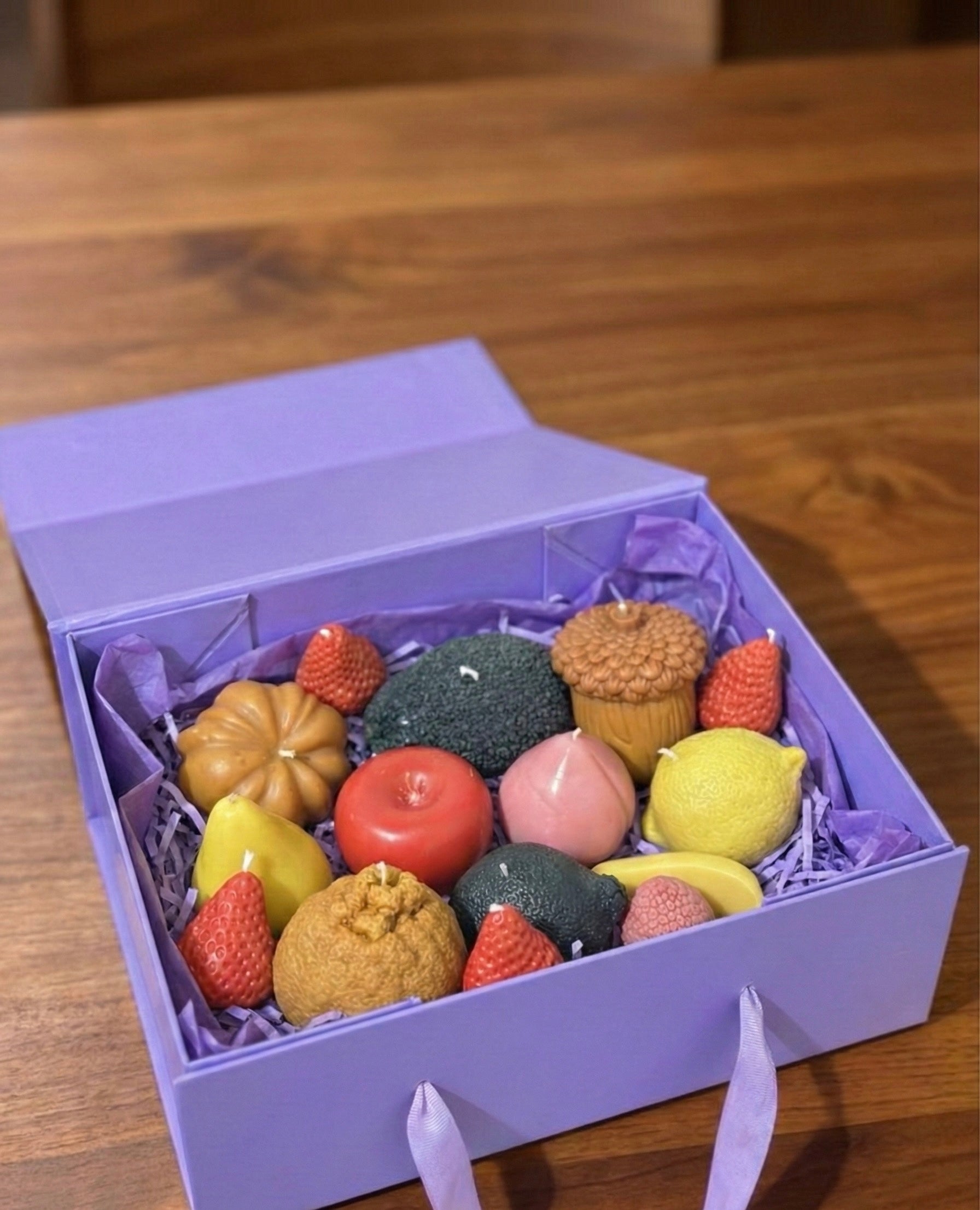 An open, light purple rigid gift box filled with purple crinkle paper. Inside, the realistic fruit candles are neatly packed and ready for gifting. The box features ribbon handles.