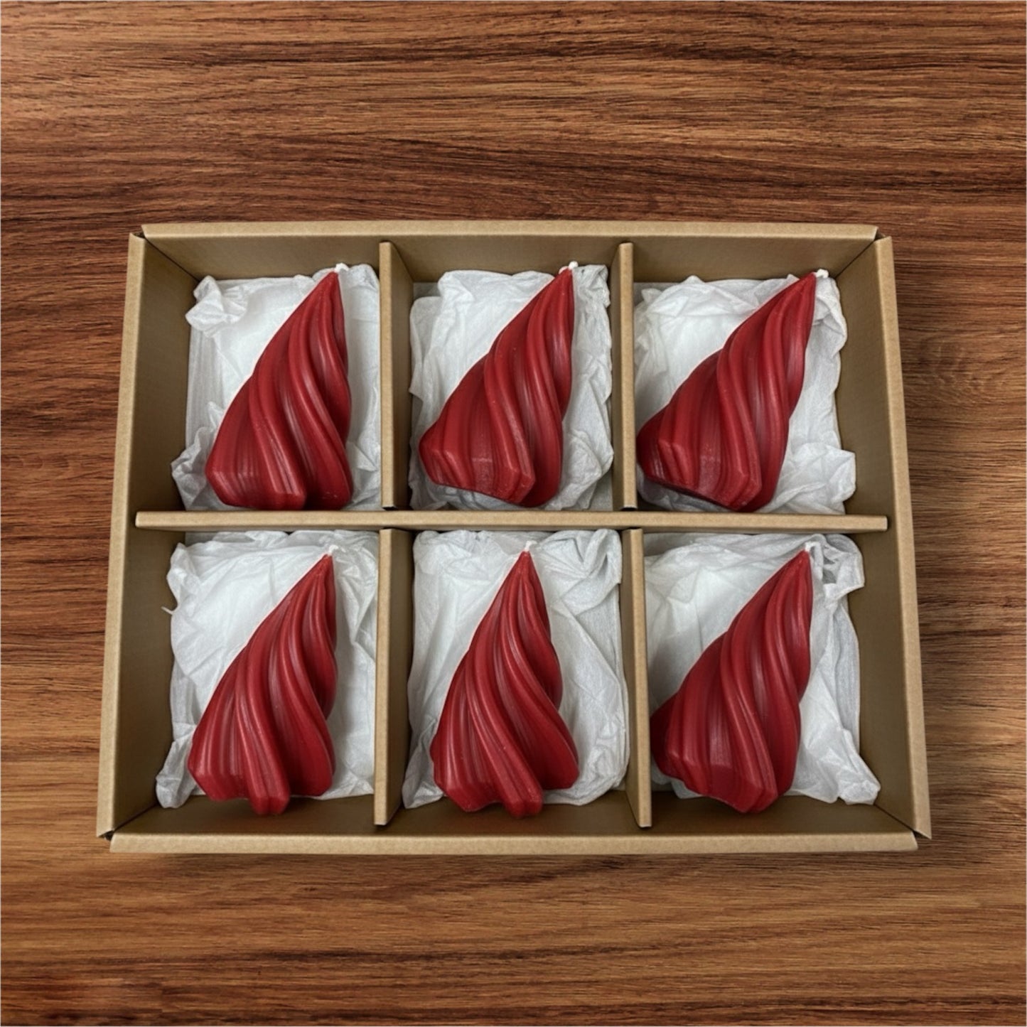 Six red, twisted/swirl sculptural candles packaged in a brown cardboard gift box with white tissue paper dividers, sitting on a wooden surface.