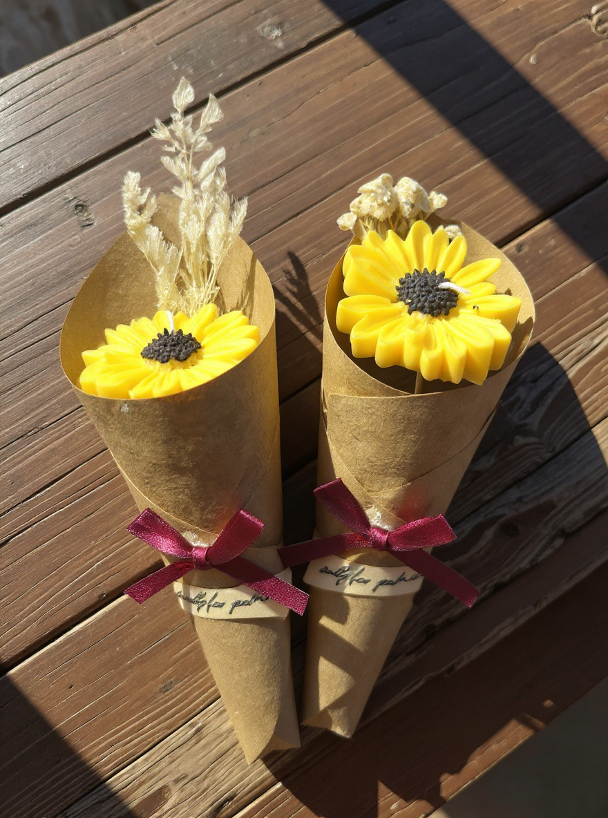 Close-up of two bright yellow sunflower-shaped candles, packaged individually in tall kraft paper cones tied with burgundy ribbons, resting on a rustic wooden table in sunlight. The packaging includes dried floral accents.