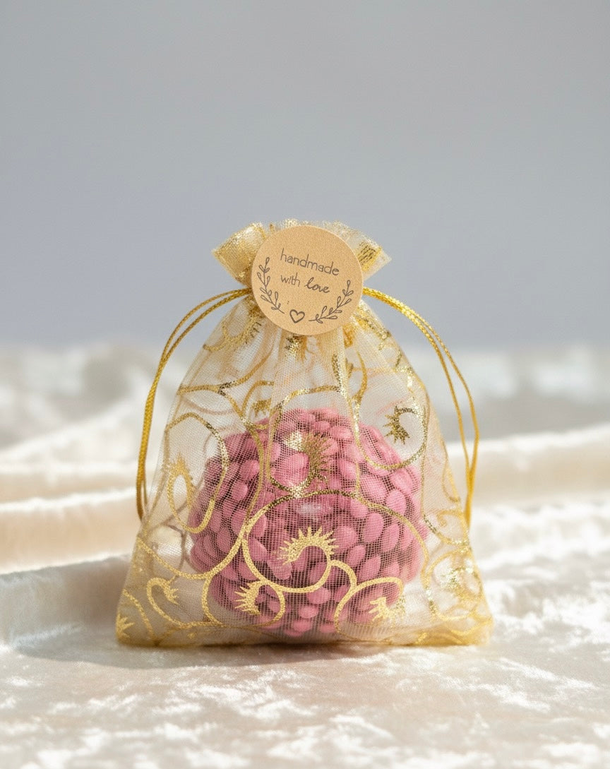 Close-up of a single pink, textured spherical candle packaged as a wedding or party favor. The candle is inside a sheer gold organza bag with filigree detailing and a round wooden tag that reads "handmade with love." The product rests on a white, velvet-textured surface.