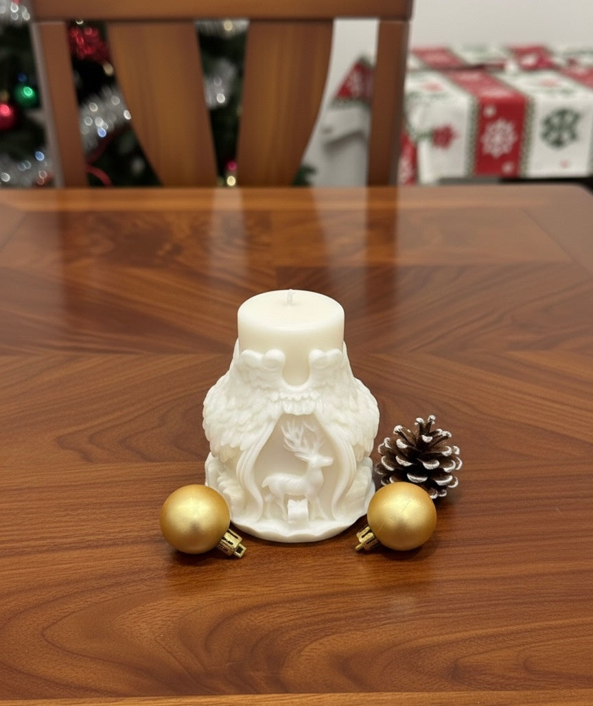 A creamy white Christmas candle featuring an intricate design of angel wings and a stag. It is displayed on a polished wooden table accompanied by two gold baubles and a frosted pinecone. A blurred dining chair and Christmas tree are visible in the background.