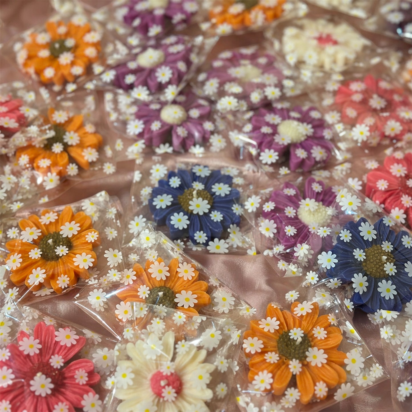 Multiple handmade orange, purple, and white daisy-shaped wax melts individually wrapped in clear cellophane bags with white daisy floral prints, arranged on a pink satin background